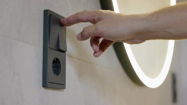 Male hand turning wall light switch on and off in modern bathroom with round illuminated mirror.
