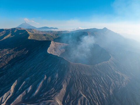 Aerial view of Mount Bromo crater with volcanic smoke, rugged terrain, and surrounding mountain peaks under a clear blue sky in Tosari, East Java, Indonesia.