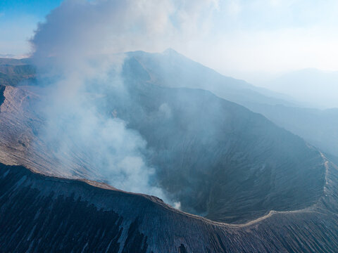 Aerial view of Mount Bromo crater with volcanic smoke rising from the caldera, rugged terrain, and hazy mountain landscape in Tosari, East Java, Indonesia.