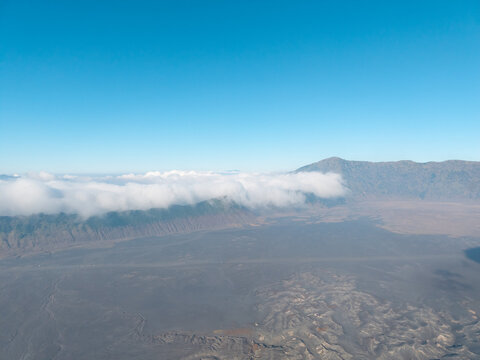 Aerial view of Mount Bromo with volcanic landscape, sand sea, and low-hanging clouds under a clear blue sky in Tosari, East Java, Indonesia.