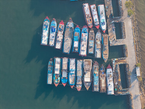 Aerial view of numerous colorful fishing boats moored in a calm harbor with clear water and a concrete pier in Probolinggo, East Java, Indonesia.