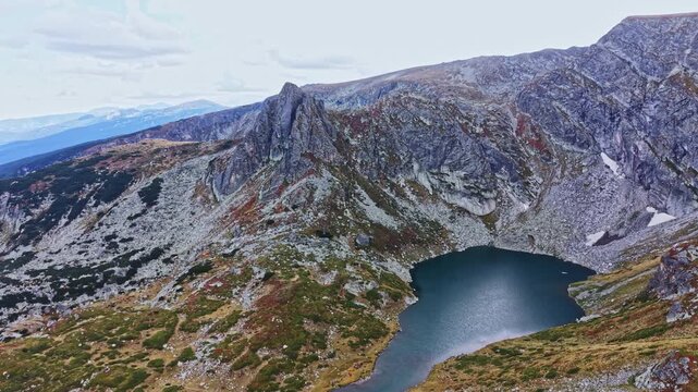 A large rocky area features a lake at its base. Mountains surround the scene, with greenery and patches of snow visible. The sky is cloudy, hinting at a cool environment.