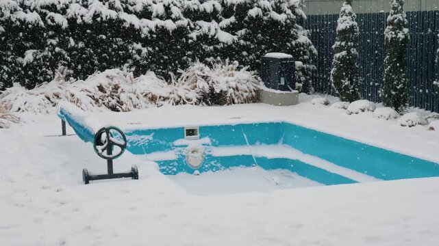 Snowfall covers backyard swimming pool with safety cover rolled up.