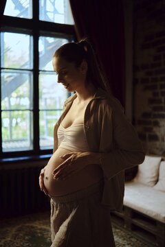 A pregnant woman stands by a large window at home, gently cradling her baby bump. The soft, warm light fills the room, creating a serene and intimate atmosphere as she enjoys this special moment