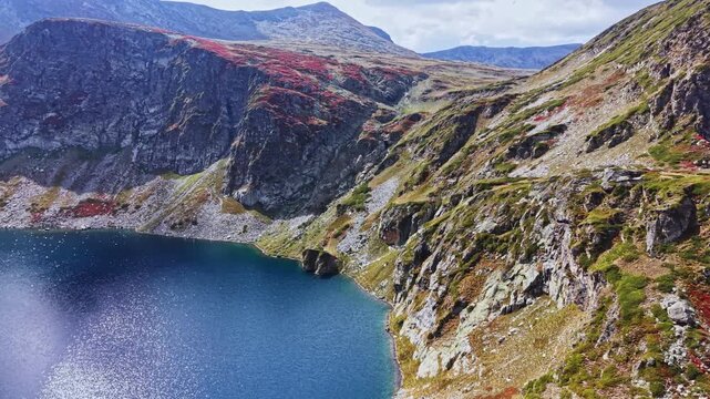 A view of mountains and a lake with clear blue water. The rocky terrain shows patches of red and green plants. The scene is bright with a blue sky above.