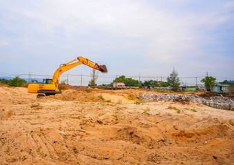 Yellow excavator on sandy construction site with raised arm, pile of waste and distant buildings under cloudy sky © MaxSafaniuk