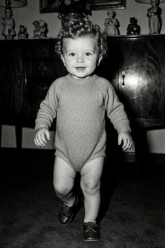Vintage black, white portrait of cute curly haired toddler boy. Small child learns to walk alone in retro home living room. Happy little boy wears knit romper, small shoes during childhood milestone.