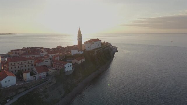 Cinematic aerial drone footage sweeping around the cliffside peninsula of Piran in Slovenia at sunset, revealing the silhouetted bell tower, Parish Church of Saint George.