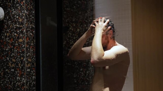 Young adult man is washing his head standing in a shower at home filmed in slow motion. Caucasian guy is taking refreshing shower massaging his head with hands. Concept of cleanliness and hygiene