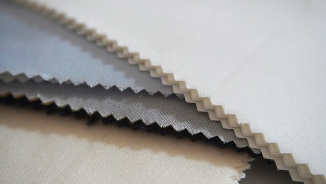Stacked fabric swatches in blue, grey, black and beige. Zigzag cut edges visible. Textile samples for fashion design and manufacturing.
