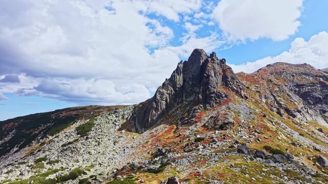 A rocky mountain peak rises above a grassy slope. The sky is filled with clouds. Colorful patches of vegetation cover the ground. This place shows nature's beauty on a clear day.