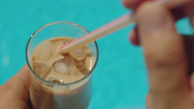 Person stirring cold iced coffee with straw near swimming pool water.