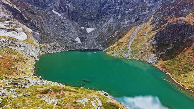 A clear blue-green lake is located in a rocky mountainous area. Surrounding hills rise steeply, and some vegetation is present. The scene shows bright daylight with no visible people.