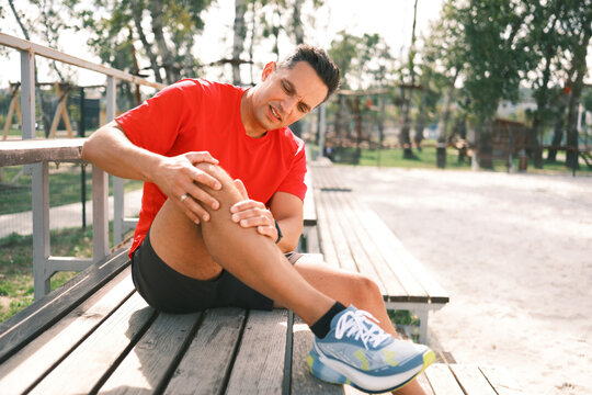 Athletic man holds his knee in pain while sitting on outdoor bleachers after running, showing a possible sports injury, discomfort and need for recovery.