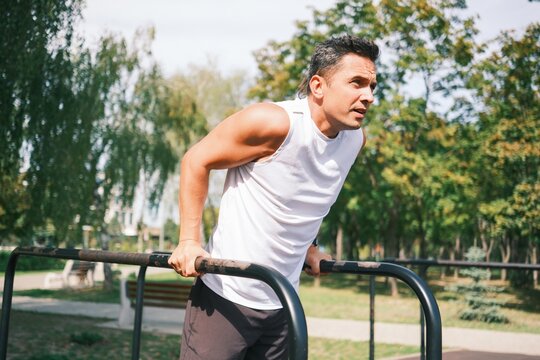 Athletic man performs dips on outdoor parallel bars in a park, building upper body strength, triceps and chest muscles during a bodyweight fitness workout.