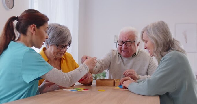 Happy senior people with dementia playing games at retirement home. Group of funny cheerful old patients sitting around table together with nurse and solving shape puzzles to improve cognitive skills