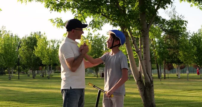 Father and son talk beside scooter in sunny city park. Parent checks child's helmet and teaches safe riding. Green summer trees create warm family recreation scene. Fatherly support builds confidence.