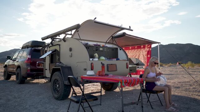 Mature woman enjoys music on a doumbek drum while sitting by her teardrop camper in the Mojave desert at sunset.