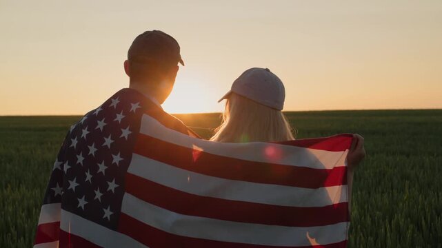 A young couple in caps with american flag on shoulders standing in green field on sunset, back view, slow motion.