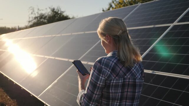 Woman engineer checks solar panels operation at sunset, using a smartphone to monitor performance and efficiency of renewable energy system.