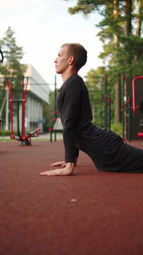 Man doing a cobra stretch outdoors. Young athlete in black athletic clothes performing a cobra pose stretch on a rubber sports ground outside. Man doing flexibility warm up routine at an outdoor gym.