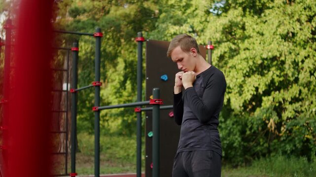 Young man in a black long-sleeve compression shirt warming up at an outdoor gym. Athlete stretching before a calisthenics training outside. Sporty man warming up outdoors.