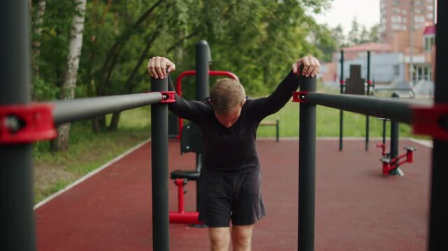 Athletic young man doing chest opening stretch at an outdoor gym. Calisthenics athlete performing a shoulder mobility stretch on parallel bars outside.