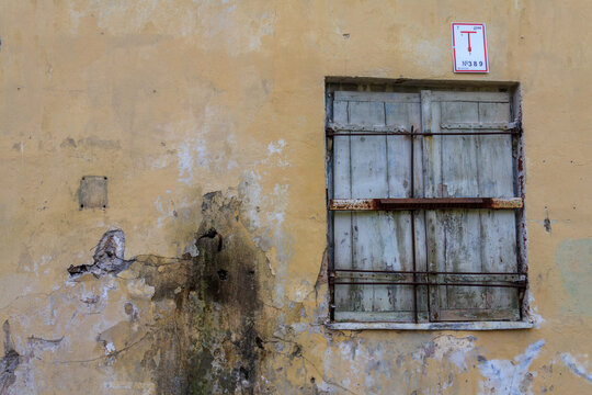 A weathered yellow plaster wall with peeling paint and a rusty barred window shuttered with old wooden panels, marked by a small numbered sign, in Tallinn, Estonia