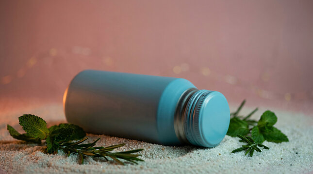 Light blue bottle with silver cap lying on sparkly sand surrounded by fresh mint and rosemary, soft pink background with bokeh lights, beverage product mockup