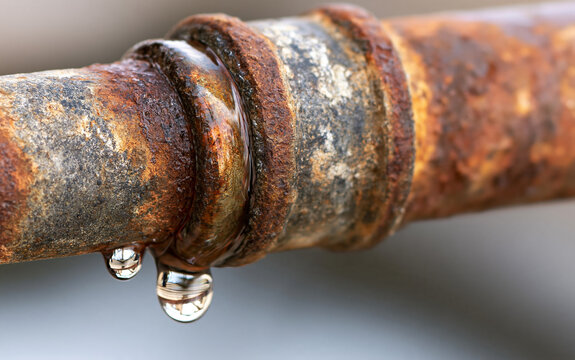 Corroded old pipe with rust texture shows dripping water drops. Metal joint connection point leaks liquid, indicating plumbing damage and system failure.