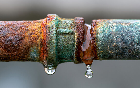 Corroded old pipe with rust texture shows dripping water drops. Metal joint connection point leaks liquid, indicating plumbing damage and system failure.