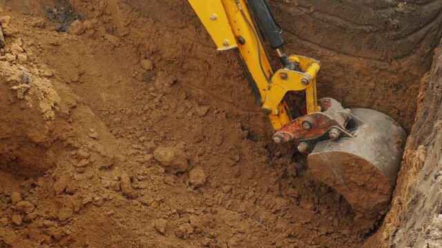 Excavator bucket digging deep pit in brown clay soil at construction site.