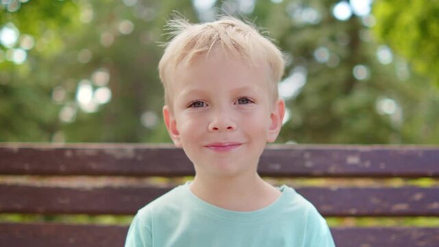 Cute little blonde boy biting an ice cream cone and smiling. Smiling kid eating a white ice cream on a bench in a bright summer park.