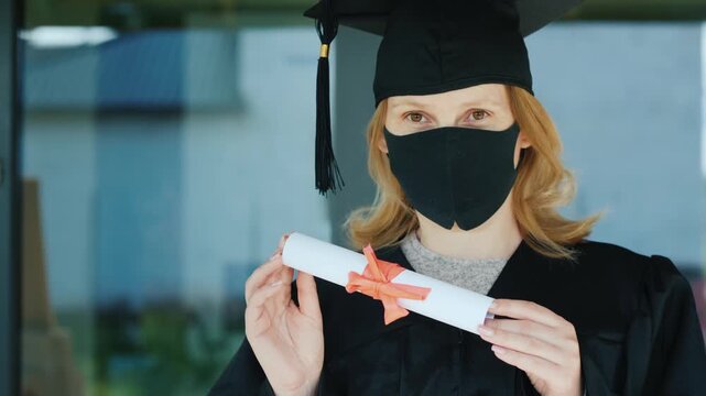 Graduate woman wearing black protective mask and academic gown holding diploma during pandemic.