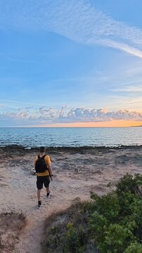 Vertical slow motion footage of a man walking from a sandy path to the shoreline at sunset, then stopping at the water's edge and looking toward the sky in Sardinia.