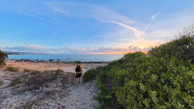 Man walks along a sandy path toward the shoreline at sunset, then stops at the water's edge and looks toward the sky, creating a peaceful coastal scene in Sardinia.