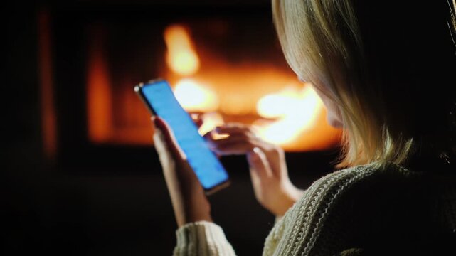 Woman using smartphone near cozy fireplace in dark room, close-up on hands and screen, warm light footage.