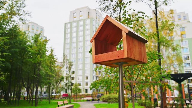 Wooden bird feeder with gabled roof on metal pole, urban park setting, leafy trees, modern apartment buildings, green foliage, residential area