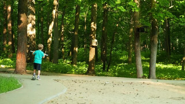 Kid riding a scooter in a forest park. Child riding a scooter outside in slow motion. Cute boy doing healthy activities outdoors.