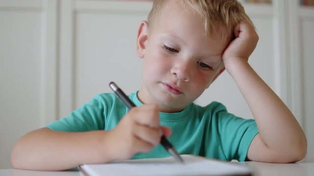 Young blonde boy writing in a notebook with a pen. Tired little boy studying in class. Sad kid writing in notebook at desk in school or kindergarten. Small student scribbling in a book.