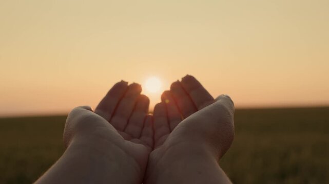 Person holding hands in cupped shape towards sunset over field.