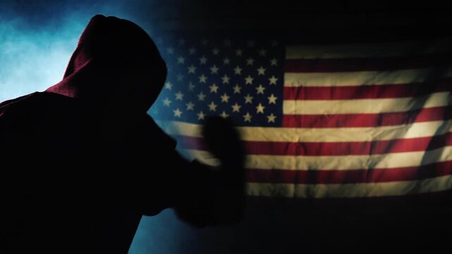 Silhouette of a man in a hoodie performing shadow boxing with an american flag background in a smoky, cinematic setting.