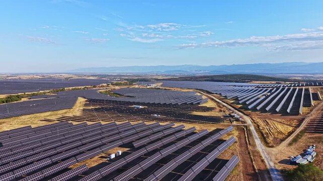 Rows of solar panels cover a large area in the countryside. The clear blue sky shows no clouds. The landscape is mostly flat with some low hills in the distance.