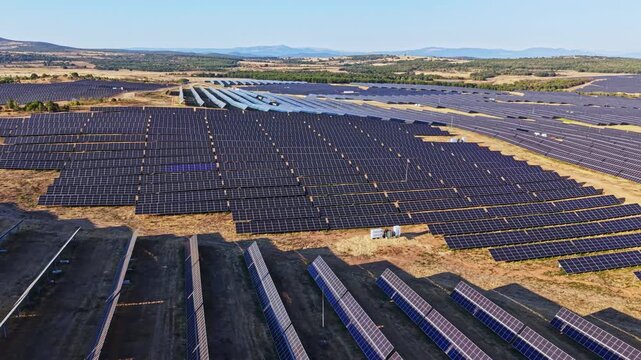 Rows of solar panels stretch across a vast field in Spain. The sun is shining, and the landscape includes mountains in the distance.