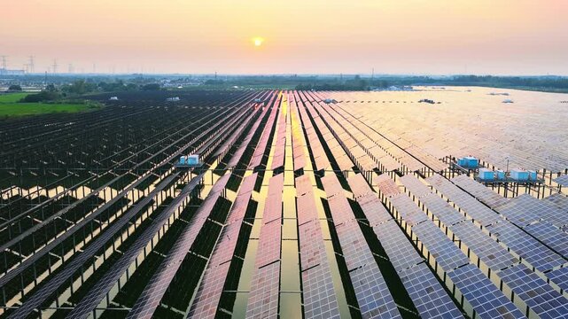Elevated view shows photovoltaic panels across a vast solar farm at sunset, catching warm light above the rural horizon.