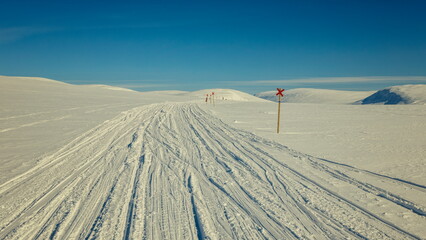 Enontekiö, Finland - 02.04.2023: Groomed snow tracks winding through vast arctic hills under a clear blue sky with red trail markers © Andrei