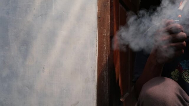 A Javanese man sits casually in front of his house, smoking a kretek clove cigarette during a quiet moment at the doorway.