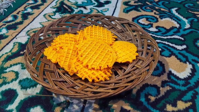 Traditional Indonesian crispy egg roll cookies or opak gambir with round waffle pattern, served on a woven bamboo plate against a patterned carpet background.