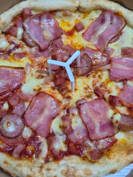 A close-up of two boxed pizzas loaded with ham, bacon, sausage, and melted cheese, each held up by a white plastic pizza saver in the center.