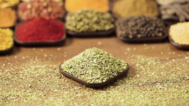 Mentha piperita or mint leaves flakes in a square bowl on a wooden table, surrounded by various spices and herbs.